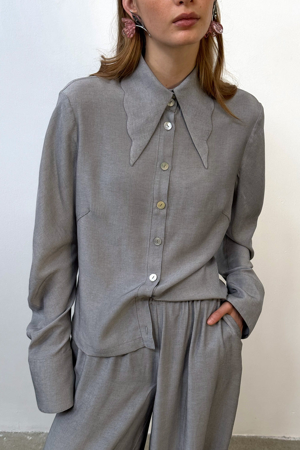 Soft grey oversized blouse with a gentle ruffled collar and relaxed silhouette, photographed against a clean neutral background